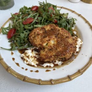 A plate with Baked Cauliflower Milanese topped with brown sauce, served over a bed of creamy spread, alongside arugula salad with halved cherry tomatoes. The gold-rimmed plate rests on a white surface.