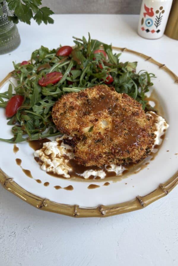 A plate with Baked Cauliflower Milanese topped with brown sauce, served over a bed of creamy spread, alongside arugula salad with halved cherry tomatoes. The gold-rimmed plate rests on a white surface.