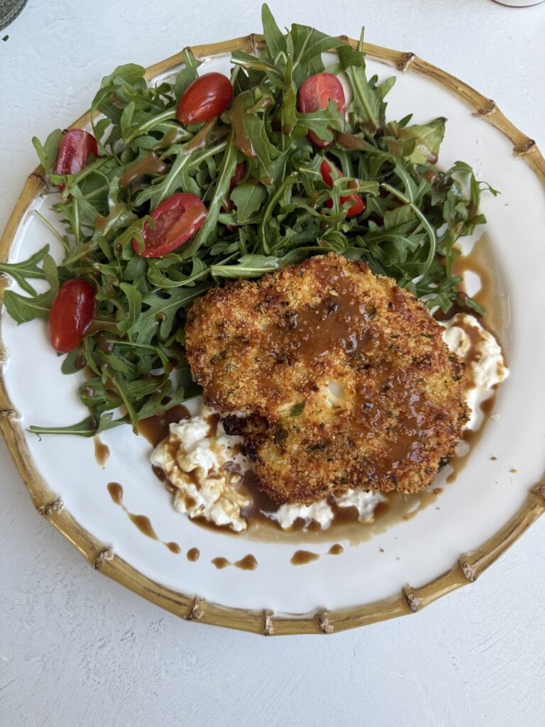 A plate with a crispy Baked Cauliflower Milanese patty on a bed of creamy cottage cheese, drizzled with brown sauce, served alongside a fresh arugula and cherry tomato salad.