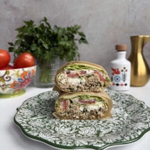 A cut Greek Salad Lettuce wrap filled with lettuce, tomato, quinoa, and feta sits on a decorative green-and-white plate. In the background are tomatoes, parsley, a patterned jar, and a gold vase on a white surface.