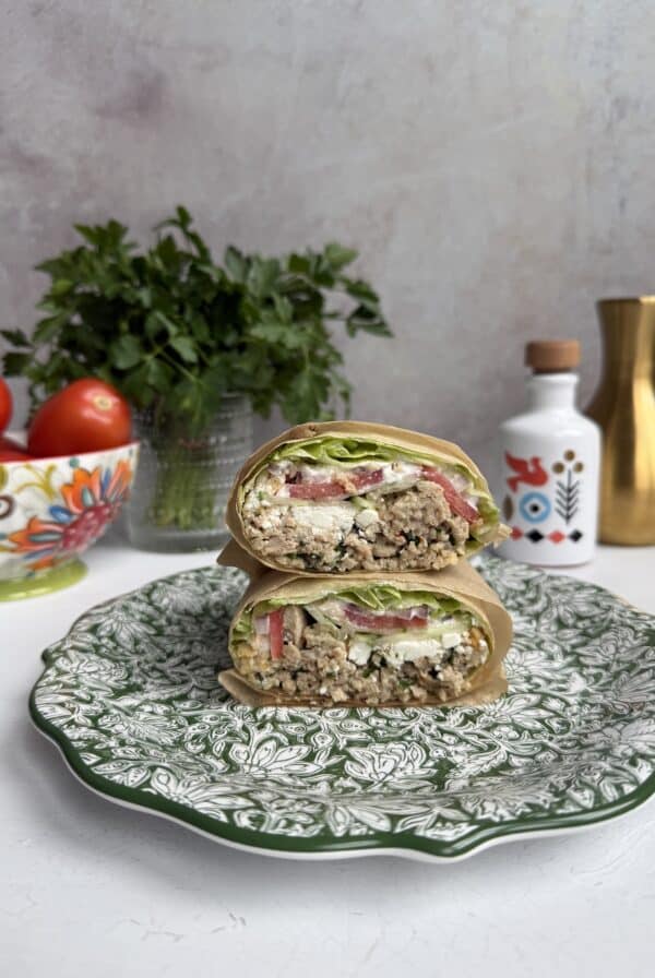 A cut Greek Salad Lettuce wrap filled with lettuce, tomato, quinoa, and feta sits on a decorative green-and-white plate. In the background are tomatoes, parsley, a patterned jar, and a gold vase on a white surface.