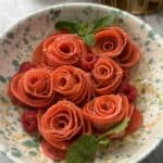A bowl with thin Rose Crepes arranged in a bowl and garnished with fresh mint leaves and raspberries, sits on a speckled ceramic plate. Lemons are visible in a basket in the background.