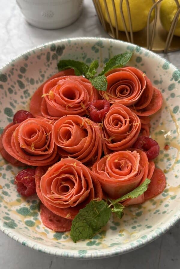 A bowl with thin Rose Crepes arranged in a bowl and garnished with fresh mint leaves and raspberries, sits on a speckled ceramic plate. Lemons are visible in a basket in the background.