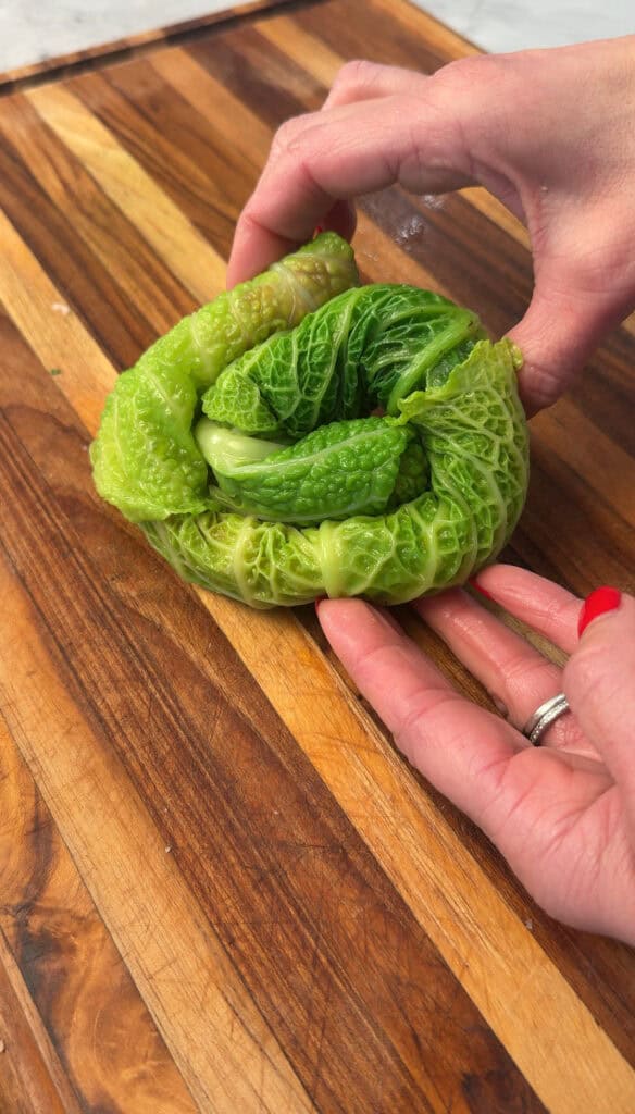 A pair of hands with red nail polish gently shape bright green cabbage leaves into a round form on a wooden cutting board.
