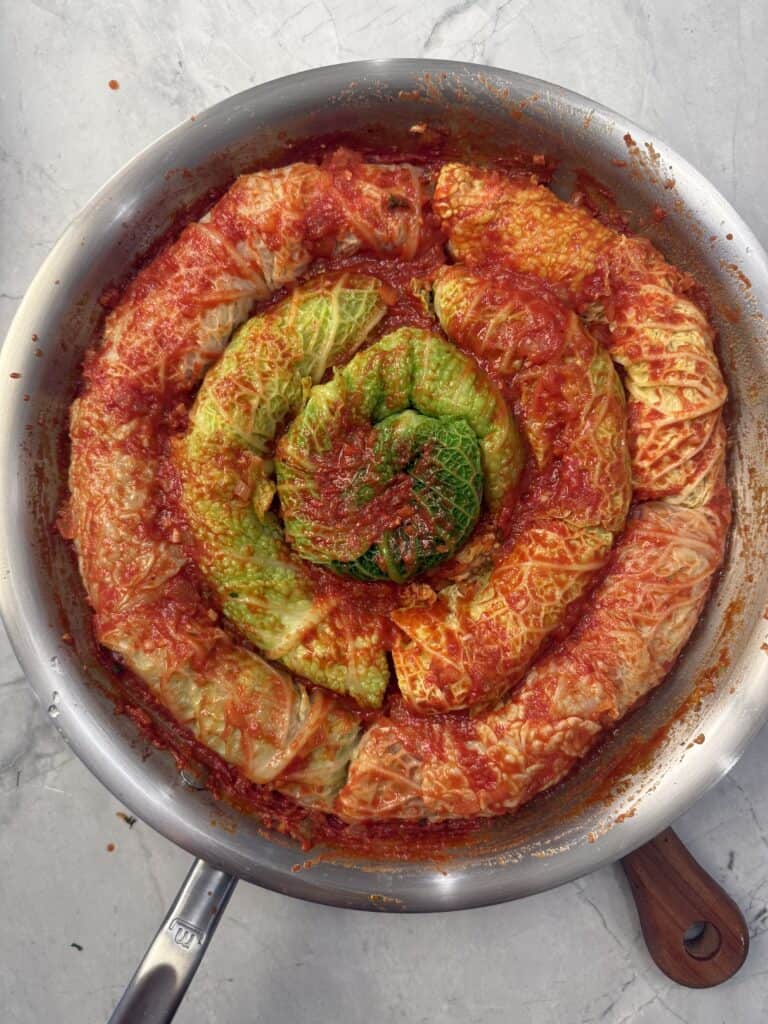 A round pan filled with cabbage rolls in tomato sauce, arranged in a spiral pattern with green cabbage leaves in the center, on a light marble surface.