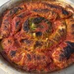 A round metal pan filled with cabbage rolls baked in tomato sauce, arranged in a spiral. The tops are browned and slightly charred, and a green leaf is visible in the center. The dish sits on a light-colored surface.