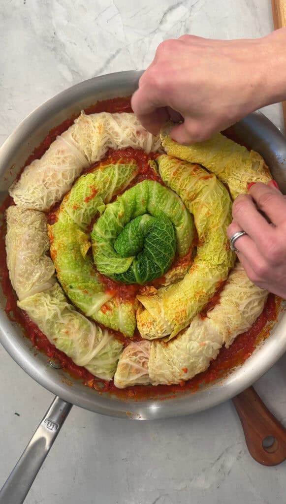 A person’s hands arrange rolled cabbage leaves in a spiral pattern in a pan filled with tomato sauce on a light countertop.