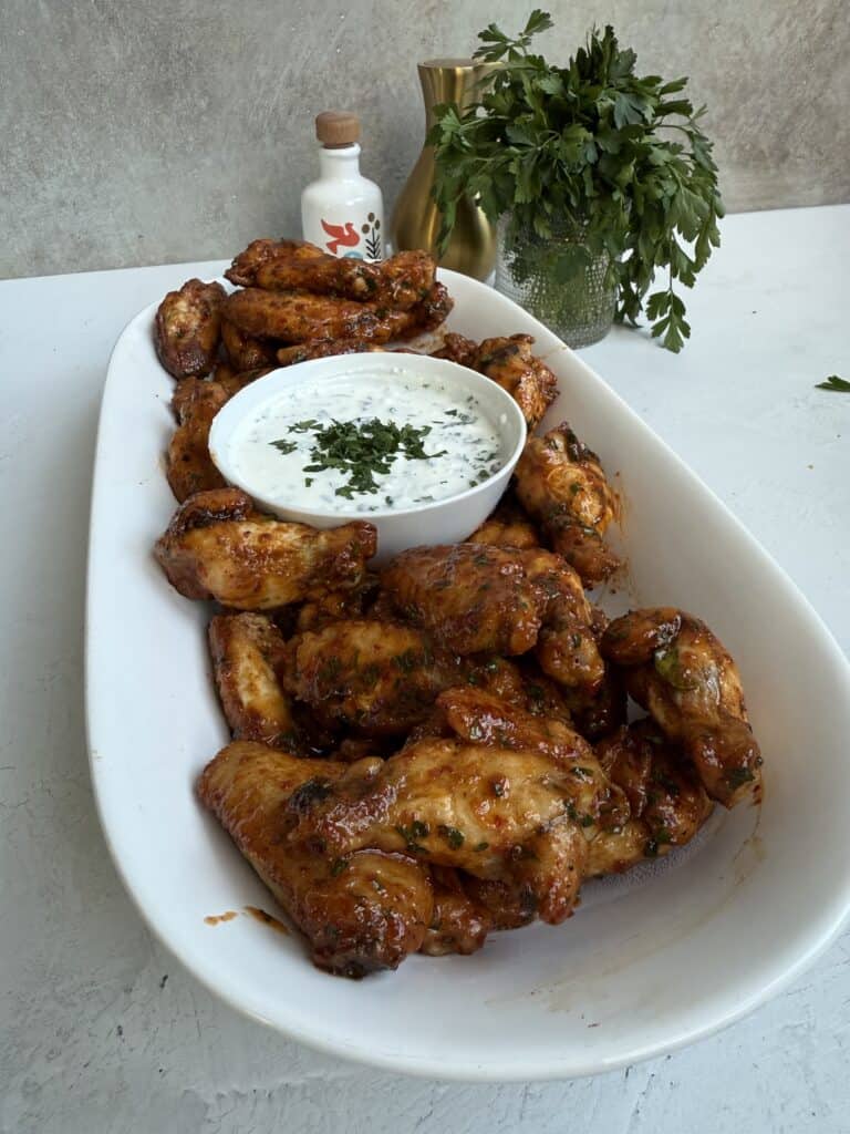 A large oval white platter filled with glazed chicken wings surrounds a bowl of creamy dip topped with herbs. Fresh parsley and a decorative bottle are in the background on a white surface.
