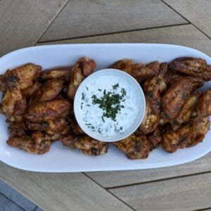 A white oval plate with a pile of easy harissa honey wings and a bowl of creamy ranch dip topped with chopped herbs, placed on a wooden table.