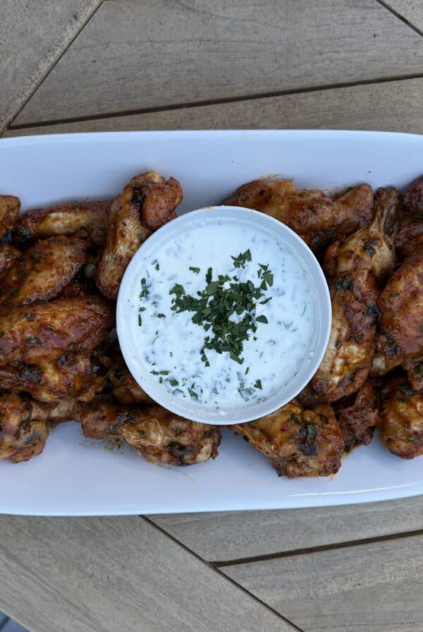 A white oval plate with a pile of easy harissa honey wings and a bowl of creamy ranch dip topped with chopped herbs, placed on a wooden table.