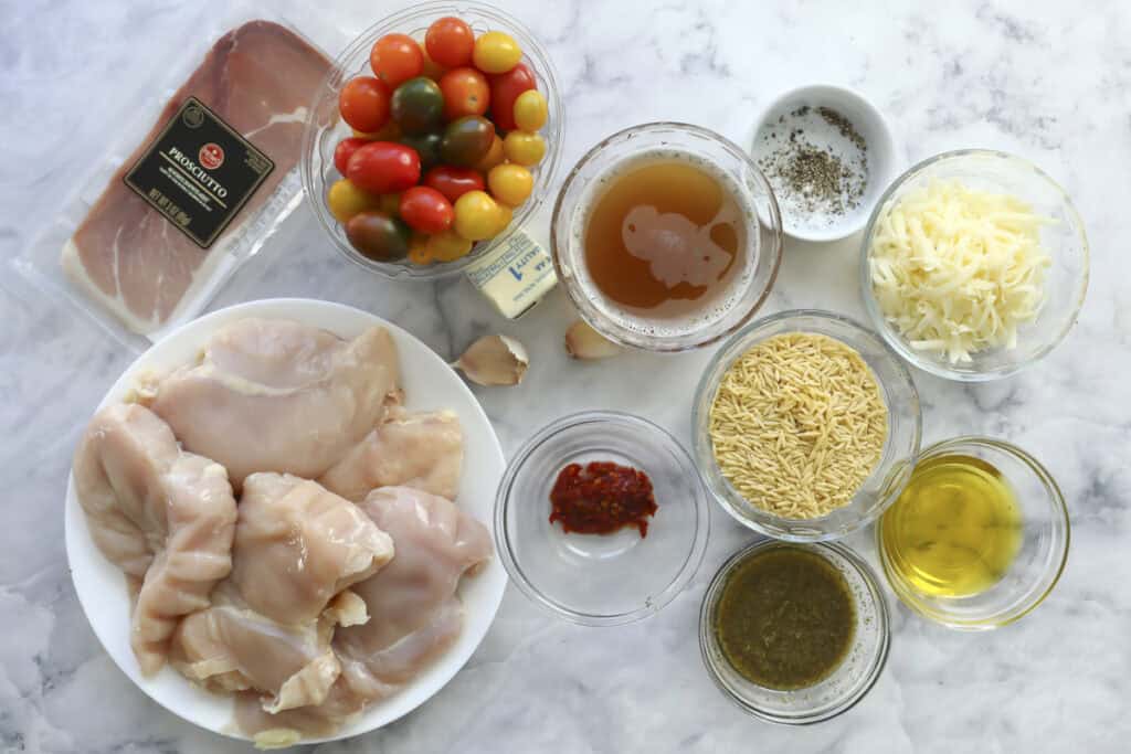 A marble counter displays the makings of a delicious Stuffed Chicken Dinner: raw chicken breasts, mixed cherry tomatoes, prosciutto, orzo pasta, shredded cheese, butter, olive oil, broth, pesto, chili paste, garlic, and seasonings in bowls.