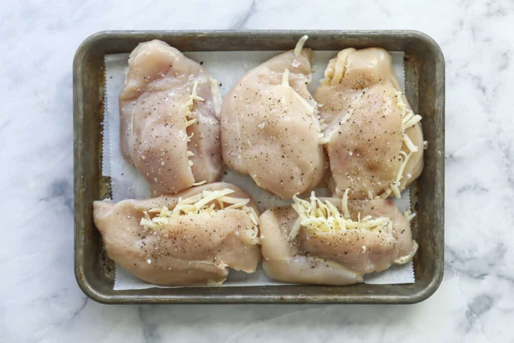 Raw chicken breasts on a baking tray, some topped with shredded cheese and all seasoned with salt and pepper, make for an easy stuffed chicken dinner on a marble surface.