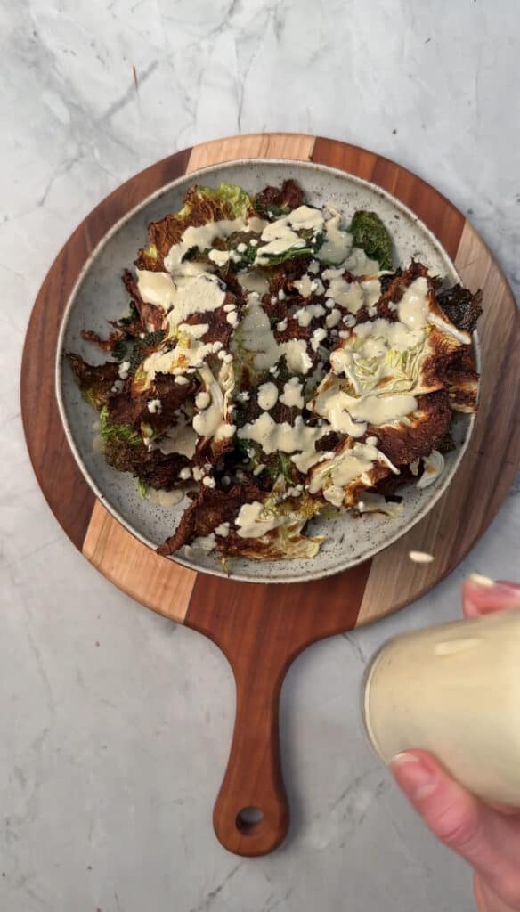 A plate of cabbage chips topped with a creamy sauce and crumbled cheese sits on a wooden serving board. A hand pours extra sauce from a glass jar onto the dish.