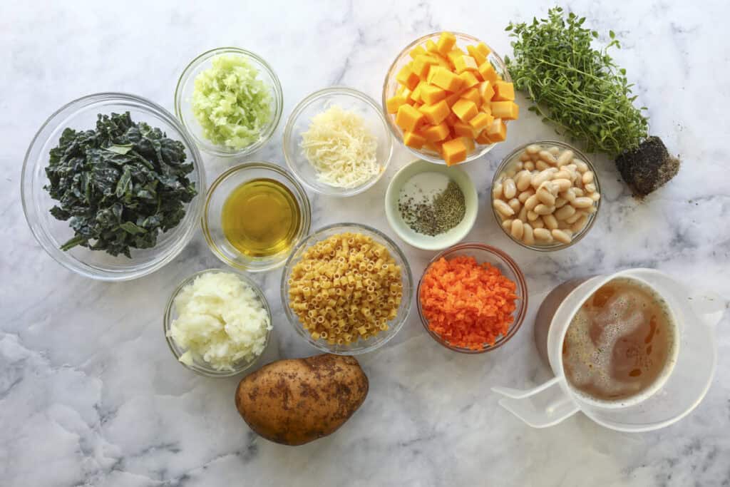 Various ingredients on a marble surface, including chopped kale, celery, carrots, onion, diced potatoes, cubed squash, white beans, olive oil, grated cheese, dry pasta, seasonings, fresh thyme, and a cup of vegetable broth.