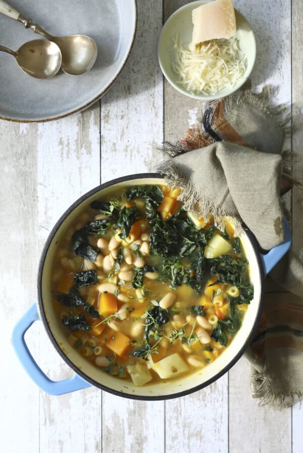 A pot of winter minestrone soup with beans, pasta, kale, and diced vegetables sits on a rustic wooden table next to a bowl of grated cheese, a chunk of Parmesan, stacked plates, and two spoons.