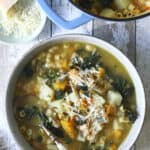 A bowl of winter minestrone soup with pasta, kale, beans, and cheese sits on a rustic wooden table. A pot of the same soup and a small dish of grated cheese are nearby. A spoon rests in the bowl.