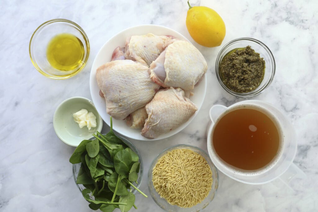 A marble surface with bowls of raw chicken thighs, olive oil, pesto, chicken broth, orzo pasta, fresh spinach, a lemon, and a piece of butter arranged neatly for meal preparation.