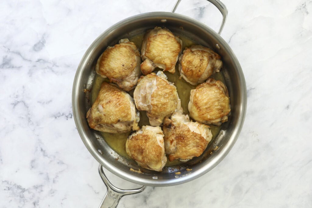 A stainless steel pan filled with eight browned chicken thighs sits on a white marble surface, with juices pooling at the bottom of the pan.