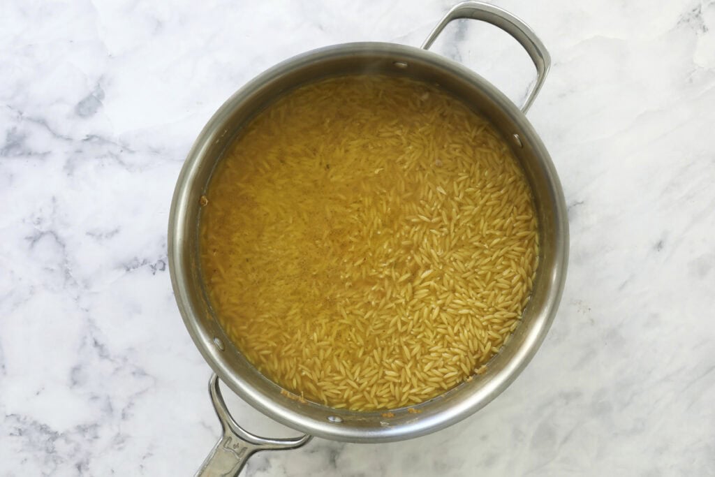 A stainless steel pot filled with orzo pasta cooking in a yellowish broth, placed on a white marble surface.