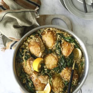 A one pan chicken & pesto dinner featuring a skillet filled with golden-brown chicken thighs, orzo pasta, spinach, and lemon wedges, placed on a light marble surface beside a stack of plates, cutlery, and a folded cloth napkin.