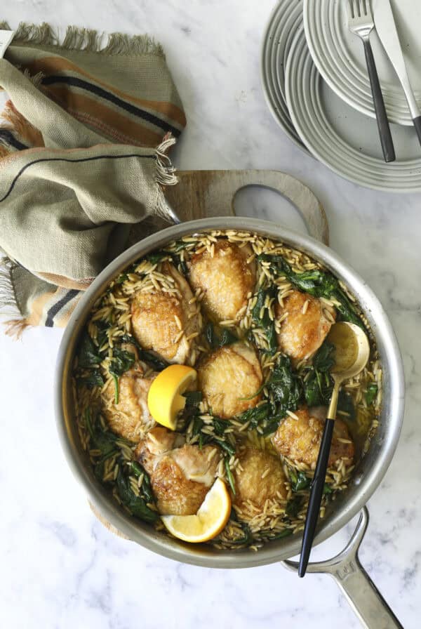 A one pan chicken & pesto dinner featuring a skillet filled with golden-brown chicken thighs, orzo pasta, spinach, and lemon wedges, placed on a light marble surface beside a stack of plates, cutlery, and a folded cloth napkin.