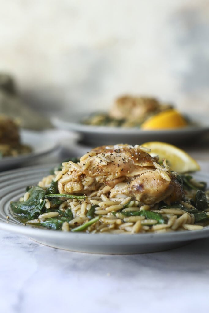 A plate of orzo pasta mixed with spinach, topped with a piece of seasoned grilled chicken, and garnished with black pepper. A lemon wedge sits beside the food. Blurred plates are visible in the background.