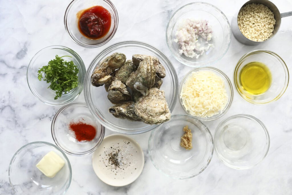 Top view of various ingredients in bowls on a marble surface for Oysters Mosca, including raw oysters, chopped parsley, shallots, grated cheese, olive oil, breadcrumbs, butter, spices, ketchup, minced garlic, salt, and pepper.