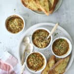 Four small ramekins filled with a baked Oysters Mosca dip, topped with breadcrumbs and herbs, are served with toasted bread slices on a round white plate. Additional bread and a pink-striped napkin are nearby. Three spoons rest on the plate.