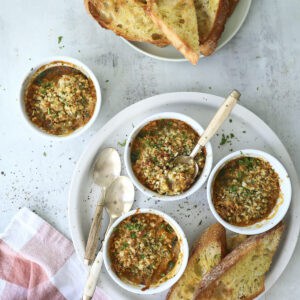 Four small ramekins filled with a baked Oysters Mosca dip, topped with breadcrumbs and herbs, are served with toasted bread slices on a round white plate. Additional bread and a pink-striped napkin are nearby. Three spoons rest on the plate.