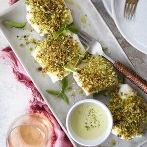A rectangular tray features Pistachio Crusted Halibut—four pieces of white fish topped with a green pistachio crust, garnished with herbs. A small bowl of creamy sauce and a glass of rosé sit nearby, plates and a fork in the background.