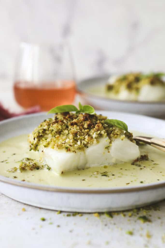 A plate with Pistachio Crusted Halibut topped with a golden, herb-infused crust, served in a creamy green sauce. A fork rests on the plate, and a glass of rosé wine is blurred in the background.