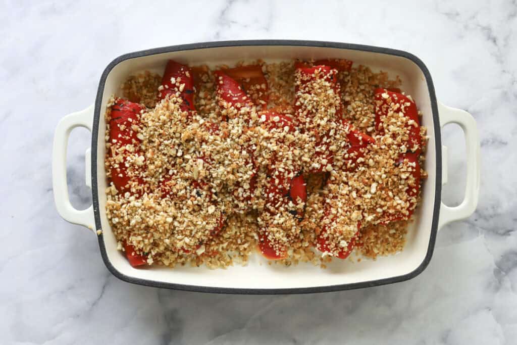 A white baking dish filled with roasted red bell peppers topped with a golden, crumbly mixture of breadcrumbs or grains, set on a marble surface.