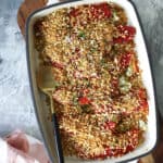 A white baking dish filled with red pepper rollatini. The red peppers are topped with golden brown breadcrumbs, garnished with herbs, and served with a gold-handled spoon. A pink checkered towel and small bowls are nearby on a gray surface.