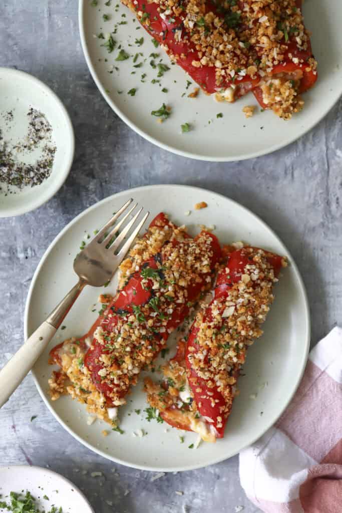 Two white plates each hold two stuffed red peppers topped with a golden breadcrumb crust and chopped herbs. A fork rests on one plate. A small dish of pepper and a pink napkin are nearby on a gray surface.