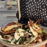A person pours a creamy dressing over a bowl of crispy cabbage chips, likely cabbage chips, in a kitchen setting. The person is wearing a dark, patterned shirt.