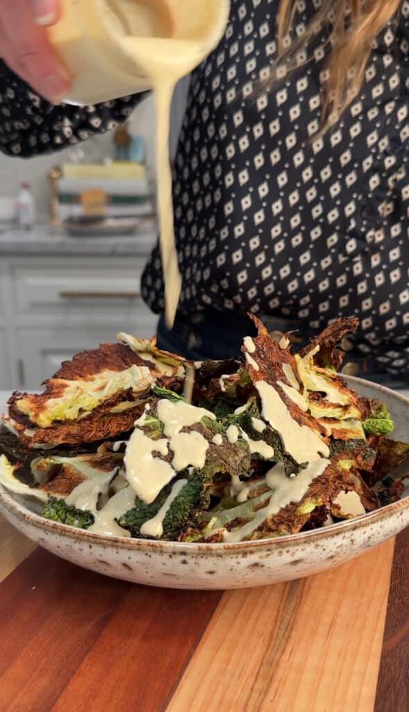 A person pours a creamy dressing over a bowl of crispy cabbage chips, likely cabbage chips, in a kitchen setting. The person is wearing a dark, patterned shirt.