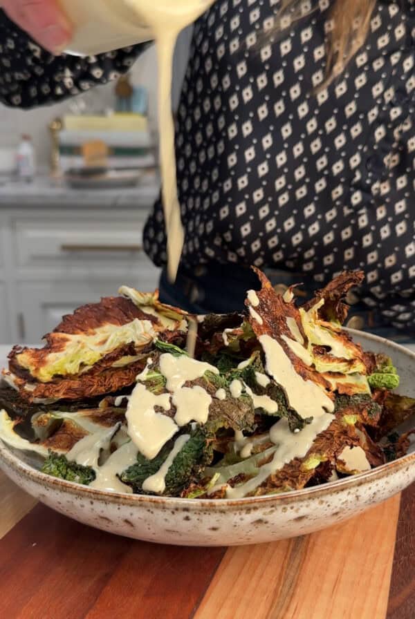 A person pours a creamy dressing over a bowl of crispy cabbage chips, likely cabbage chips, in a kitchen setting. The person is wearing a dark, patterned shirt.