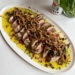 Sliced seared tuna, perfect for a 20 Minute Tuna Steak Dinner, topped with crispy fried onions, chopped herbs, and diced red peppers on an oval white plate with yellow sauce and parsley. A vase with greenery is in the background.