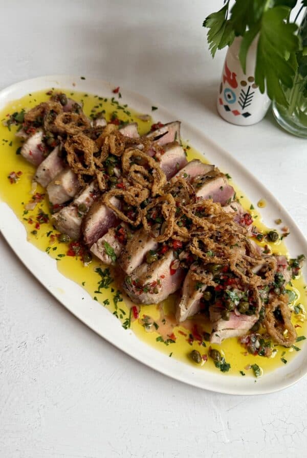 Sliced seared tuna, perfect for a 20 Minute Tuna Steak Dinner, topped with crispy fried onions, chopped herbs, and diced red peppers on an oval white plate with yellow sauce and parsley. A vase with greenery is in the background.