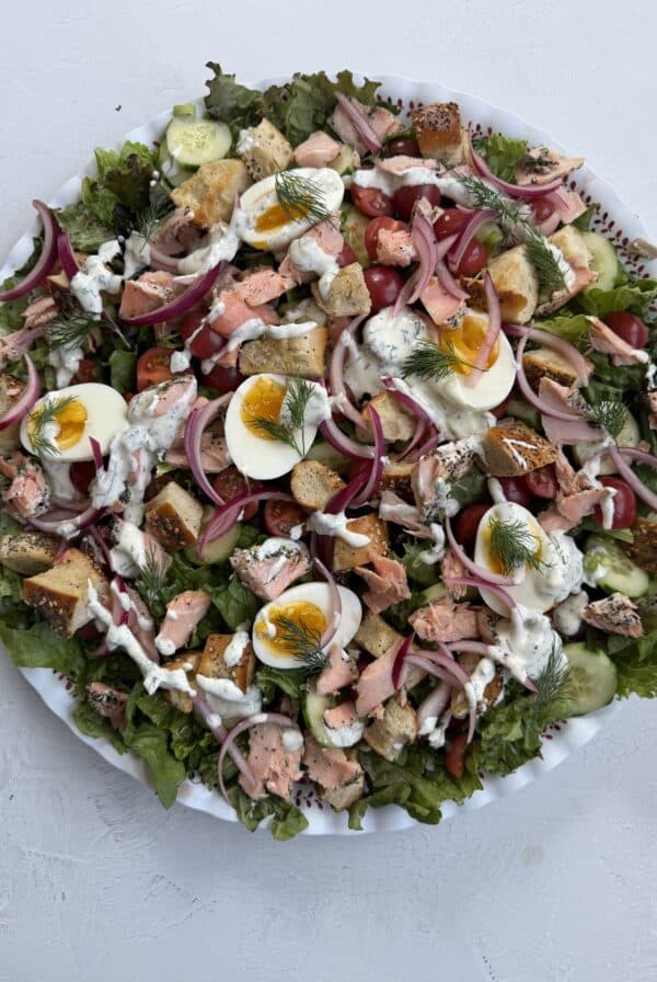 A large Everything Bagel Salmon Salad on a white plate, topped with lettuce, cucumbers, tomatoes, red onions, hard-boiled eggs, chunks of salmon, croutons, dill, and a drizzle of creamy dressing.