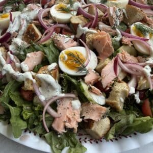 An Everything Bagel Salmon Salad topped with leafy greens, hard-boiled egg halves, flaked salmon, red onion slices, croutons, and creamy dressing, garnished with fresh dill on a white plate.