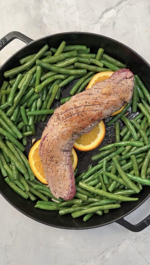 A cast iron skillet with green beans, orange slices, and an uncooked Orange Glazed Pork Tenderloin, all beautifully arranged on a light-colored surface.