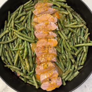 Orange Glazed Pork Tenderloin is sliced and arranged with orange sauce in the center of a skillet, surrounded by cooked green beans on a light-colored surface.