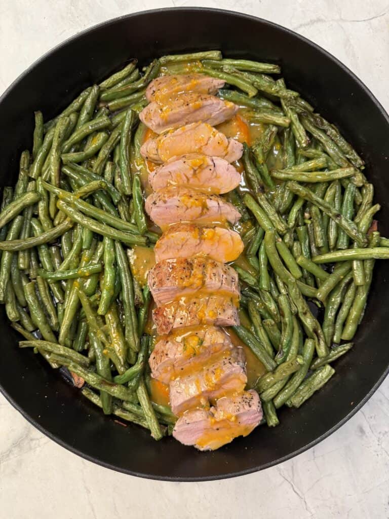 Orange Glazed Pork Tenderloin is sliced and arranged with orange sauce in the center of a skillet, surrounded by cooked green beans on a light-colored surface.