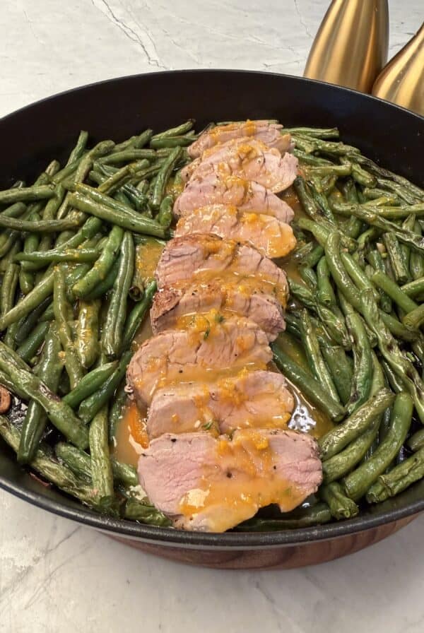 A skillet filled with roasted green beans surrounds slices of Orange Glazed Pork Tenderloin, each piece topped with a light orange glaze. Two gold decorative bottles are partially visible in the background.