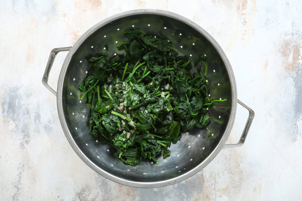 A metal colander filled with cooked spinach and chopped onions, often used in a classic Pasqualina, rests on a light-colored, textured surface.