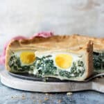 A slice of Pasqualina pie with a golden crust, filled with spinach, creamy cheese, and a whole boiled egg, displayed on a round wooden cutting board.