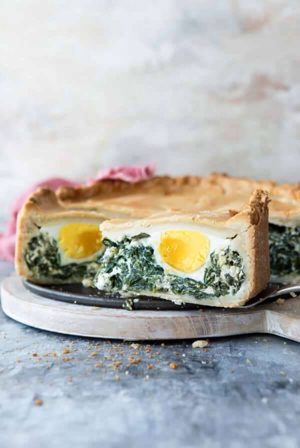 A slice of Pasqualina pie with a golden crust, filled with spinach, creamy cheese, and a whole boiled egg, displayed on a round wooden cutting board.