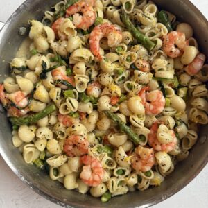 A skillet filled with shell pasta, shrimp, asparagus, and yellow squash, garnished with black pepper and herbs, on a white surface.