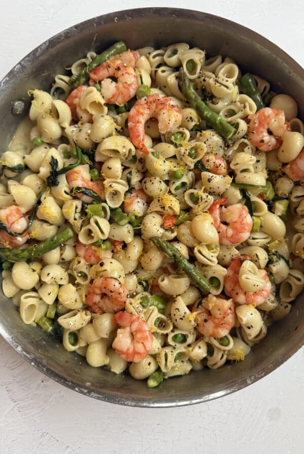 A skillet filled with shell pasta, shrimp, asparagus, and yellow squash, garnished with black pepper and herbs, on a white surface.